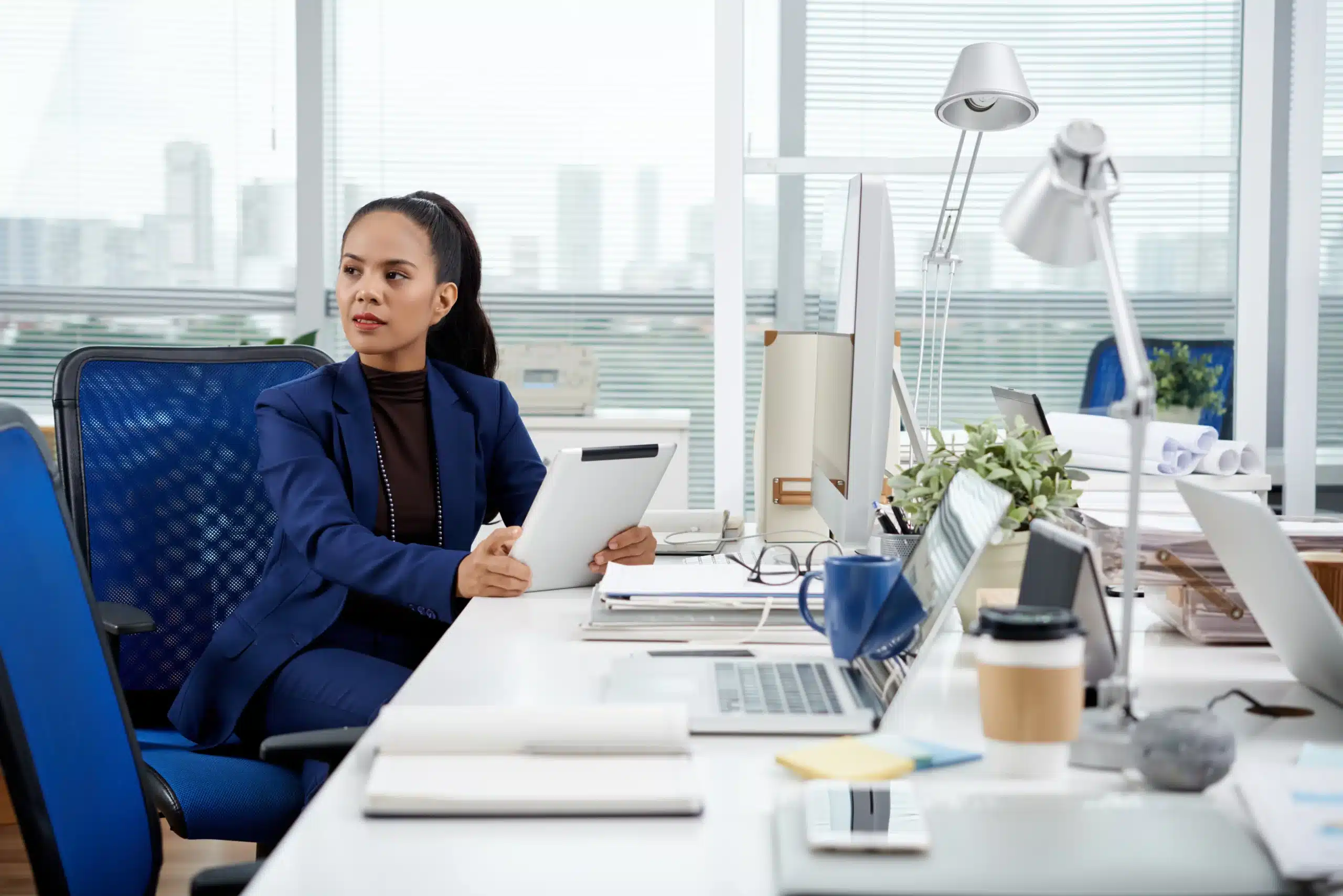 Femme professionnelle en costume bleu dans un bureau moderne, tenant une tablette, illustrant le quotidien de travailler dans l’IA.