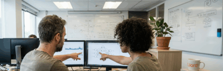 Deux collègues analysent des données sur des écrans d'ordinateur dans un bureau, avec des schémas liés à l'intelligence artificielle.