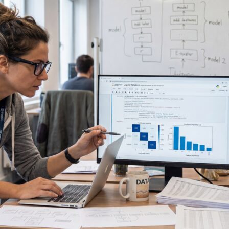 Femme travaillant sur un ordinateur portable avec deux écrans affichant des graphiques et des données dans un bureau moderne.