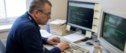 Un programmeur écrivant du code COBOL sur deux écrans d'ordinateur, entouré de livres et d'une tasse de café.