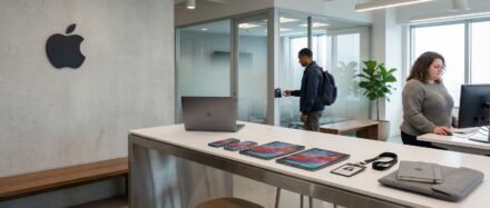 Intérieur d'un bureau moderne mettant en avant des produits Apple, avec une personne travaillant sur un ordinateur et une autre en train de discuter en arrière-plan.