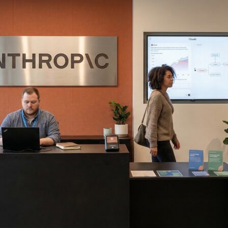 Espace d'accueil d'Anthropic montrant un homme travaillant à un bureau et une femme passant, avec, au mur, un écran interactif affichant des visualisations de données.