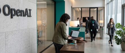 Une personne travaillant sur un ordinateur portable dans les locaux d'OpenAI, des collègues qui circulent en arrière-plan, illustrant l'innovation et la collaboration.