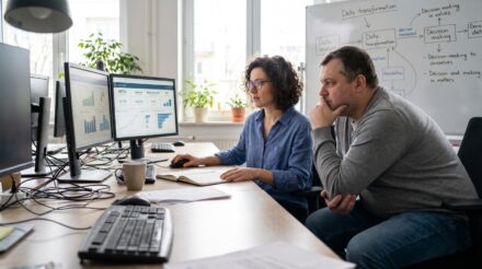 Deux professionnels analysant des données sur plusieurs écrans d'ordinateur dans un environnement de bureau.