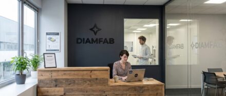 Área de recepción de Diamfab con una mujer trabajando en un ordenador portátil y un hombre en segundo plano leyendo documentos, que representa la innovación en tecnología de baterías nucleares.