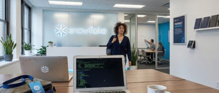 Eine Büroszene, in der eine Frau sich einem Schreibtisch mit einem Laptop nähert, auf dem Code sichtbar ist, und die Snowflake-Marke in einem modernen Arbeitsumfeld präsentiert wird.